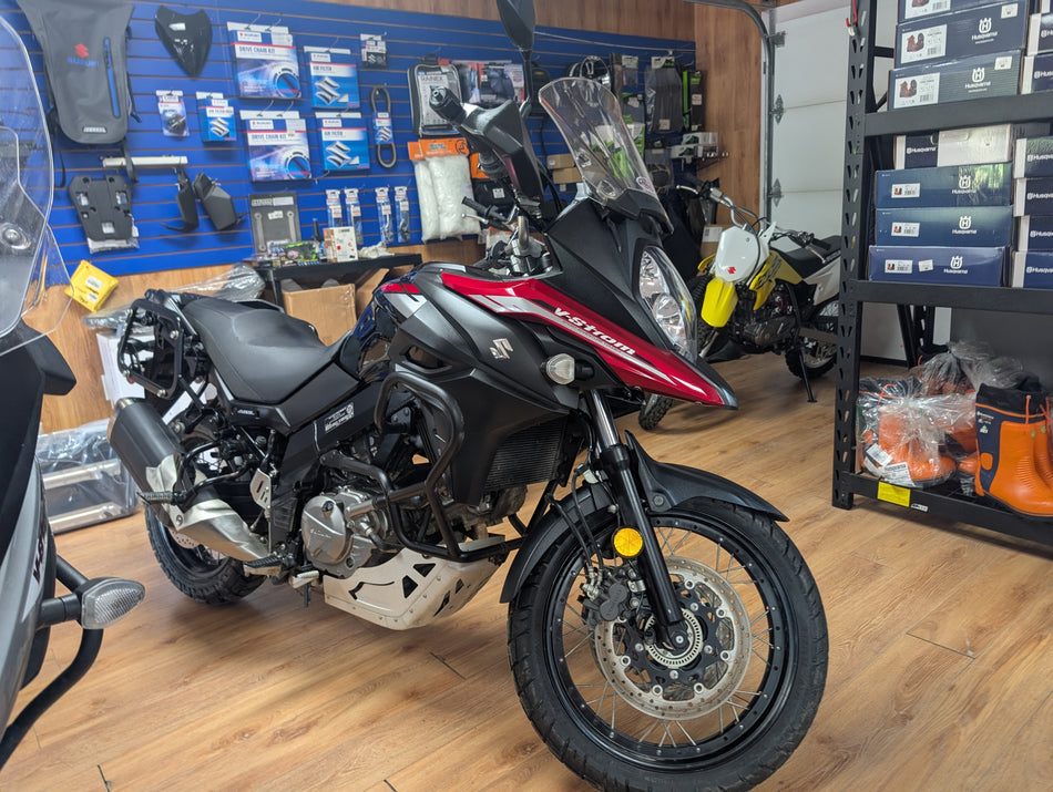 A black and red Suzuki Adventure motorcycle is parked inside a motorcycle workshop with wood floors, a blue product display wall, and storage shelves in the background.