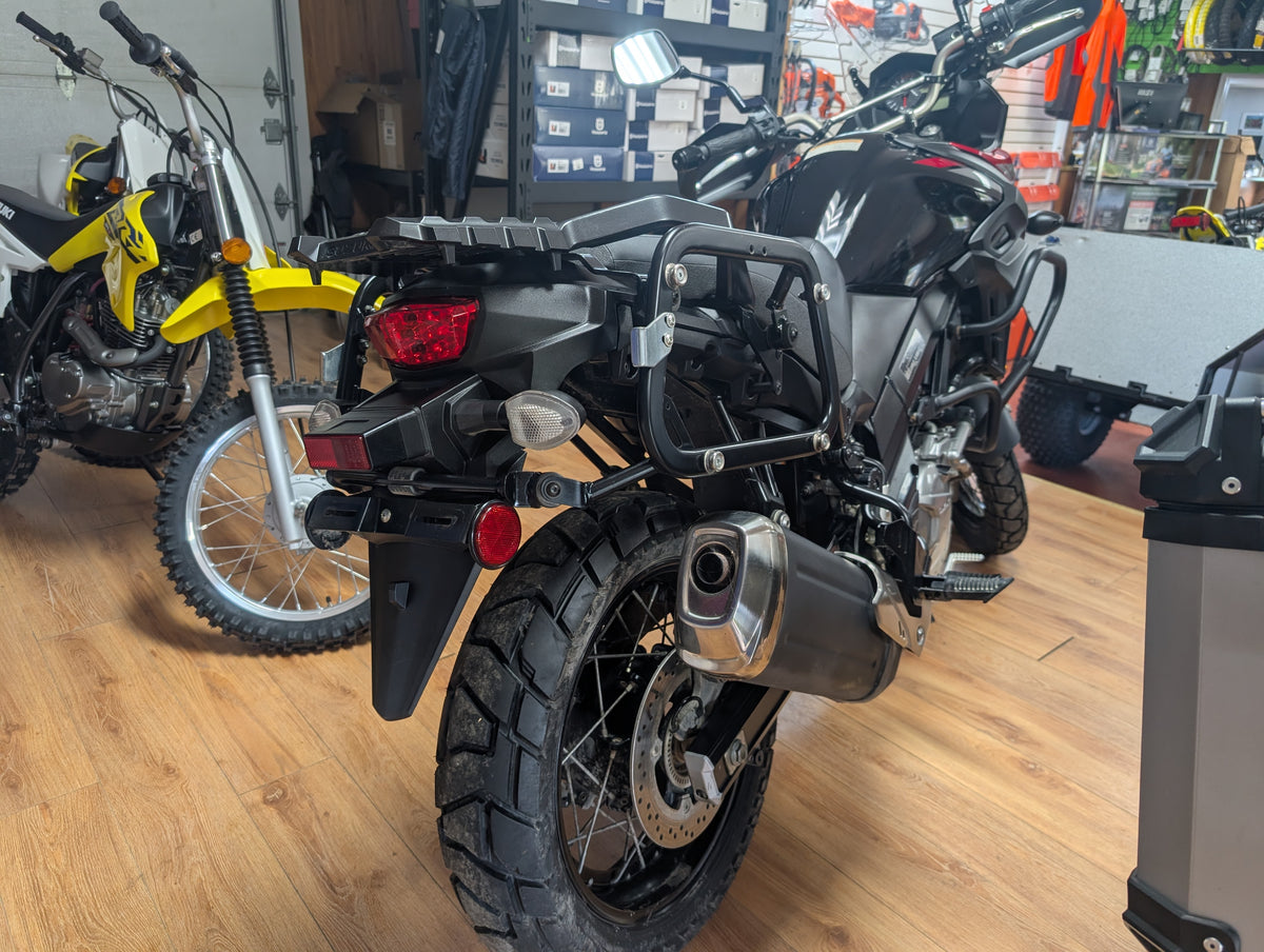A black dual-sport motorcycle with a rear luggage rack and protective crash bars, photographed inside a motorcycle dealership showroom with other bikes in the background.