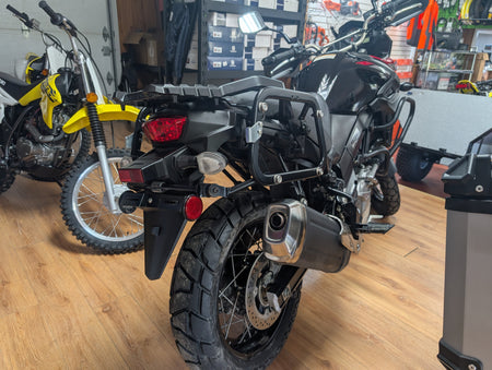 A black dual-sport motorcycle with a rear luggage rack and protective crash bars, photographed inside a motorcycle dealership showroom with other bikes in the background.
