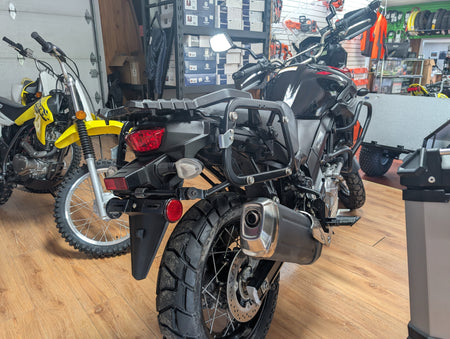 A black dual-sport motorcycle with protective crash bars and an exhaust system, photographed in a motorcycle shop with other bikes and storage shelves in the background.